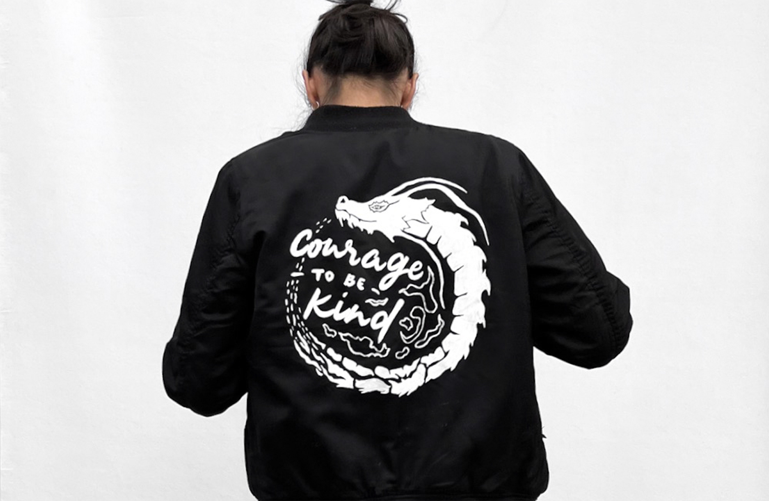 Black bomber jacket with a white Courage TO BE Kind dragon circle graphic, worn from behind
