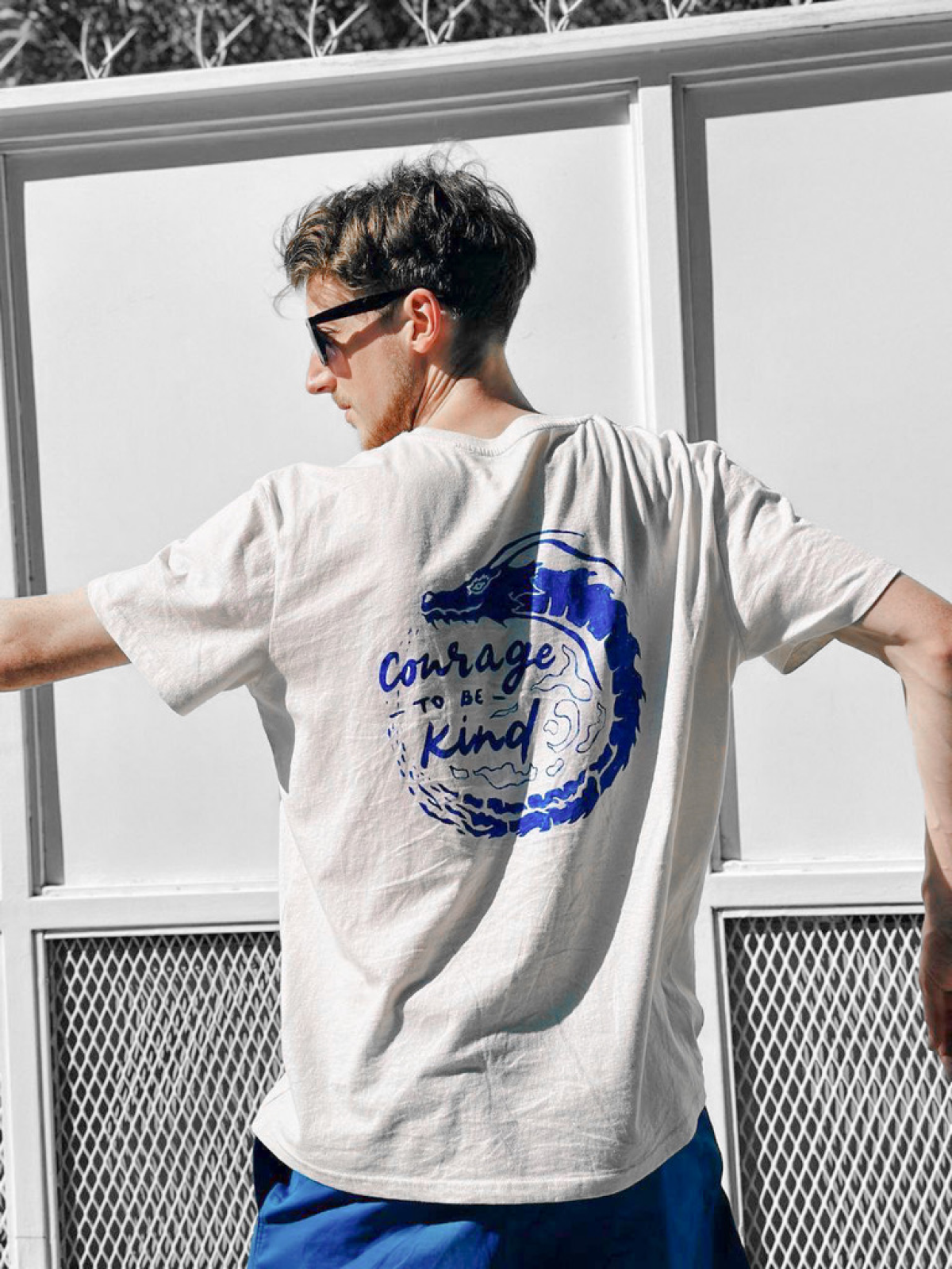 White t-shirt with a blue Courage TO BE Kind dragon design, worn outdoors from behind