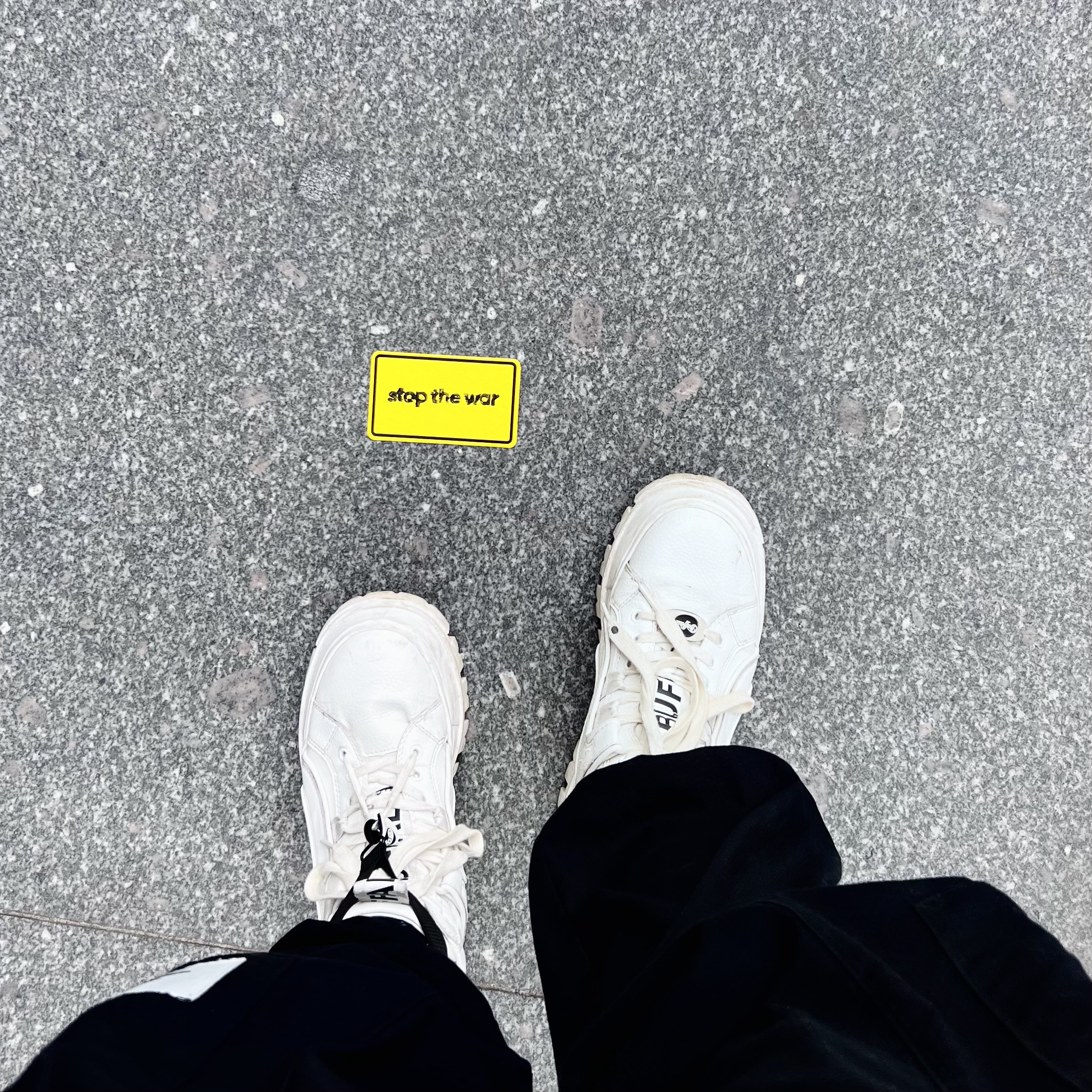 Yellow stop the war sticker on pavement photographed from above with white sneakers